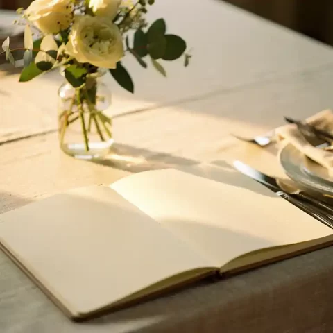 Opengeslagen notitieboek met handgeschreven checklist op een linnen gedekte tafel, omringd door servet, couvert en bloemenvaasje in warm middaglicht.