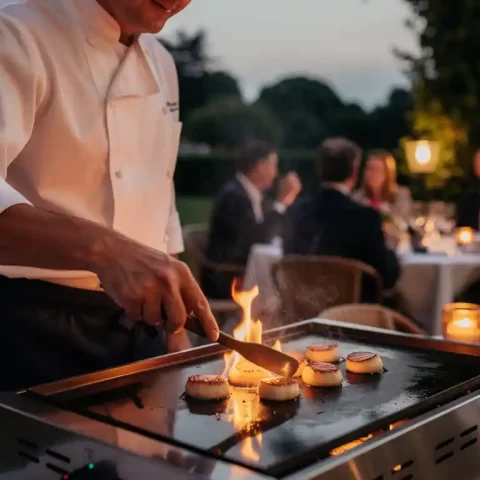 Chef in wit jasje scheurt coquilles op een plancha grill tijdens een elegant buitenevenement, met vlammen en warme kaarslichtsfeer.