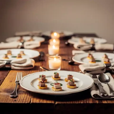 Elegante cateringtafel met witte porseleinen borden, bitterhapjes, linnen servetten en kaarslicht op rustiek houten oppervlak.