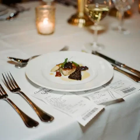 Cateringnota gedeeltelijk verborgen onder een elegant bord op een gedekte evenementstafel met zilverwerk, wijnglas en kaarslicht.