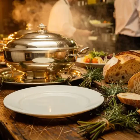 Luxe zilveren serveerschalen naast een eenvoudig bord op een rustieke houten tafel, versierd met verse kruiden, ambachtelijk brood en delicate garnering.