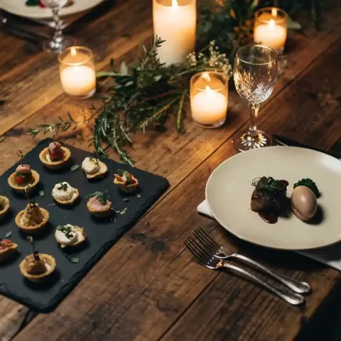 Rustieke houten tafel met borrelhapjes op leisteen borden naast een driegangendiner, verlicht door kaarsen met gouden gloed.