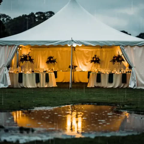 Witte evenemententent met gouden binnenverlichting, gedekte cateringtafels en regendruppels op nat gras op de voorgrond.