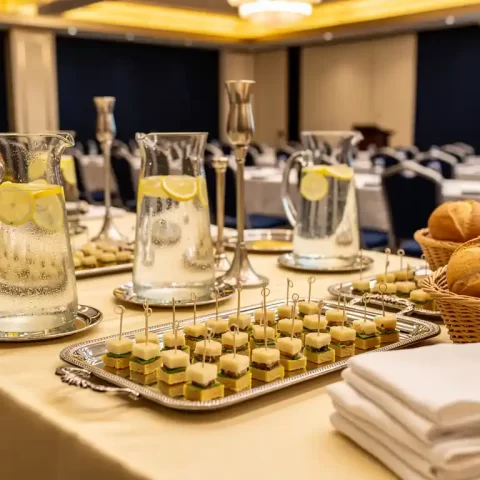 Luxe cateringtafel met zilveren schalen hapjes, glazen waterkannen met citroen en gevouwen linnen servetten in een moderne conferentiezaal.