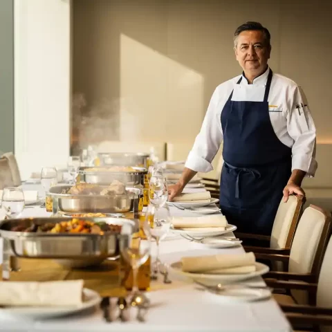 Cateringmanager in koksuniform bij lange gedekte banquettafel in moderne bedrijfsrestaurant, dampende gerechten in gouden serveerschalen.
