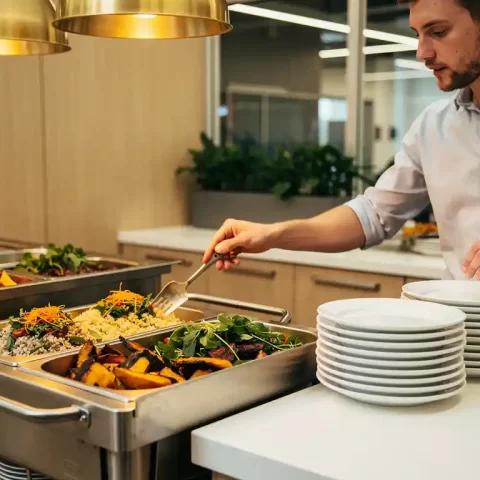 Medewerker serveert zichzelf bij een moderne bedrijfslunch met kleurrijke warme gerechten op een strak aanrecht.