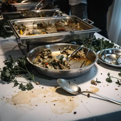 Halfleeg catering buffet met verwelkte garnering, bijna lege schalen en een vergeten opscheplepel op een gevlekt tafelkleed.