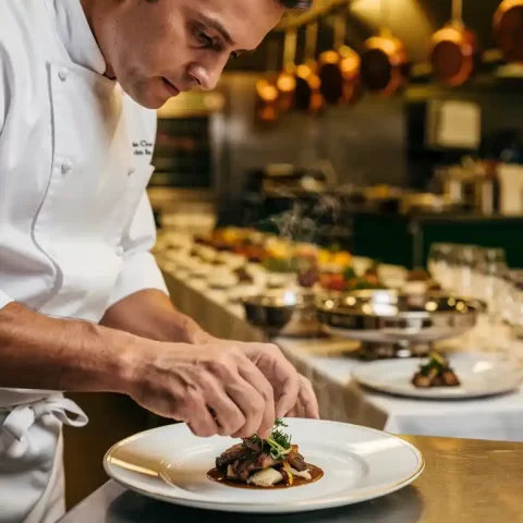 Chef in wit uniform plaatst met precisie een elegant gerecht op, warme keukenomgeving met catering opstelling op de achtergrond.