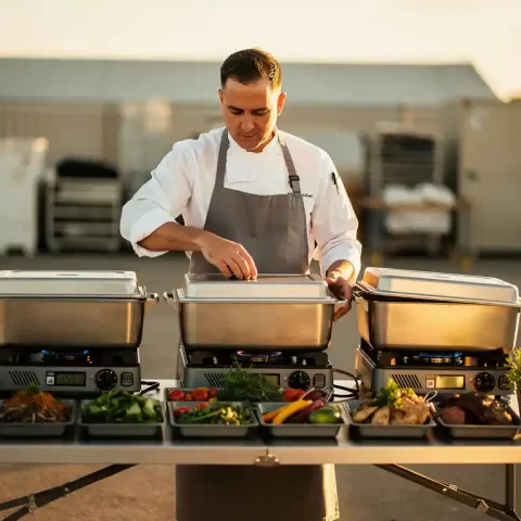 Cateringchef pakt geïsoleerde transportcontainers uit op een klaptafel bij een buitenevenement, met draagbare branders en mise en place.