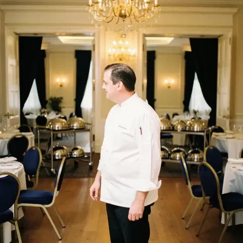 Cateringmanager in wit koksjas inspecteert elegante evenementenlocatie met zilveren trolleys, serveerdomes en tafels gedekt met wit linnen.